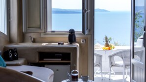 A room with a kitchenette, a window with a view of the sea, and a table with chairs outside. - La Perla Villas and Suites - Adults Only (Santorini)