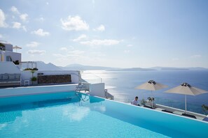 A poolside area with a view of the sea, a white building, and two umbrellas. - La Perla Villas and Suites - Adults Only (Santorini)