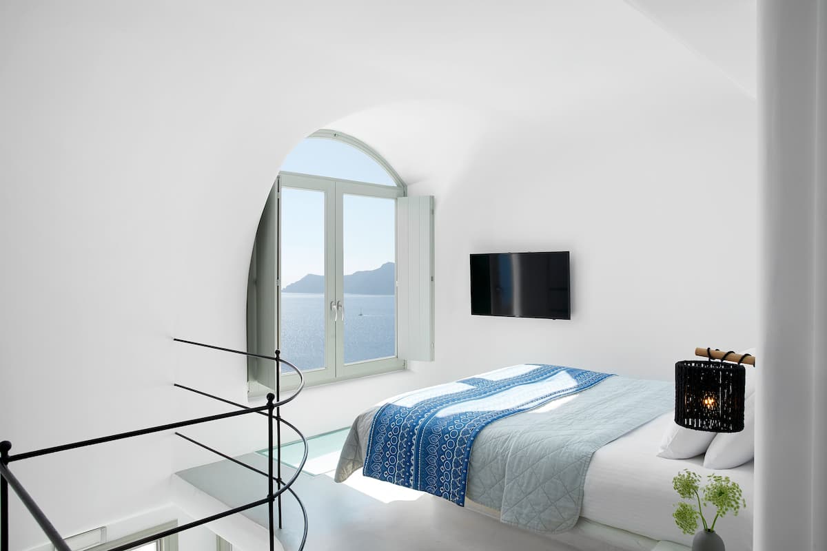 Een moderne slaapkamer met een groot rondboogvenster, een bed met een blauw-wit gebeddeken, een zwarte tv aan de muur en een zwarte lamp op een nachtkastje.