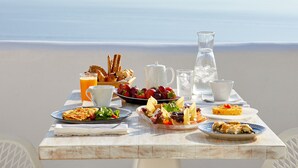 A breakfast table with various dishes, a pitcher, and a cup, set against a beach backdrop. - La Perla Villas and Suites - Adults Only (Santorini)