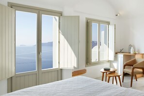 Habitació amb llit, escriptori, cadira i grans finestres que donen vistes al mar.