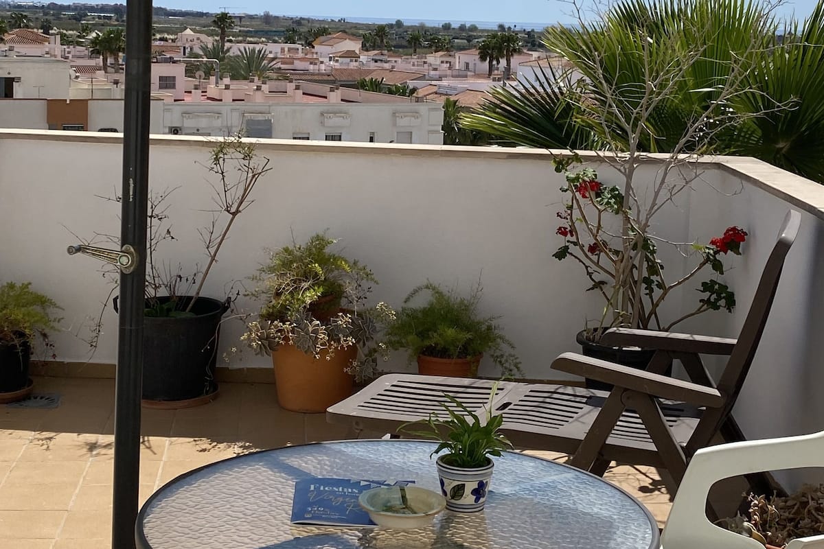 A round table with a glass top, surrounded by chairs, under a yellow umbrella on a balcony with potted plants and a view of buildings.