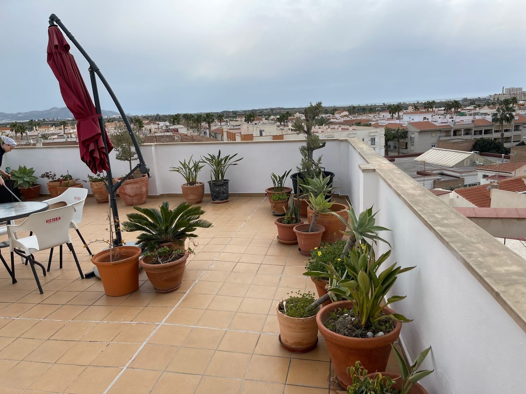 A rooftop terrace with potted plants, a red umbrella, and a table with chairs.