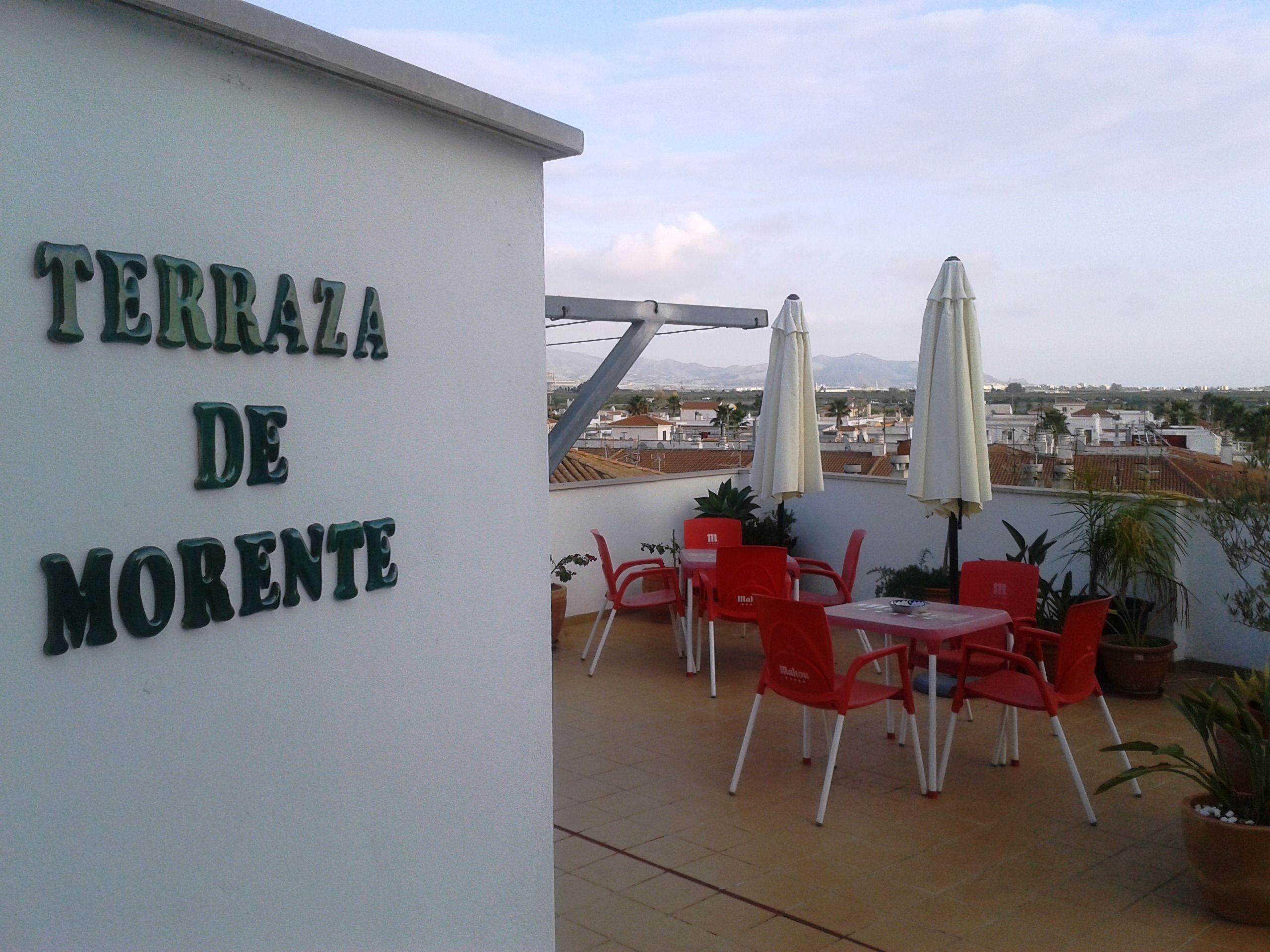 Una terraza con un cartel que dice "TERRAZA DE MORENTE". Hay sillas rojas y sombrillas blancas en la terraza.