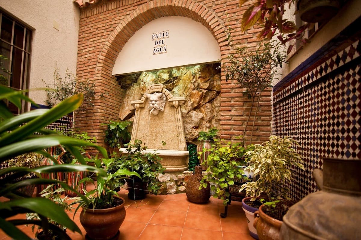 A courtyard with a stone fountain, potted plants, and a tiled wall with a geometric pattern.