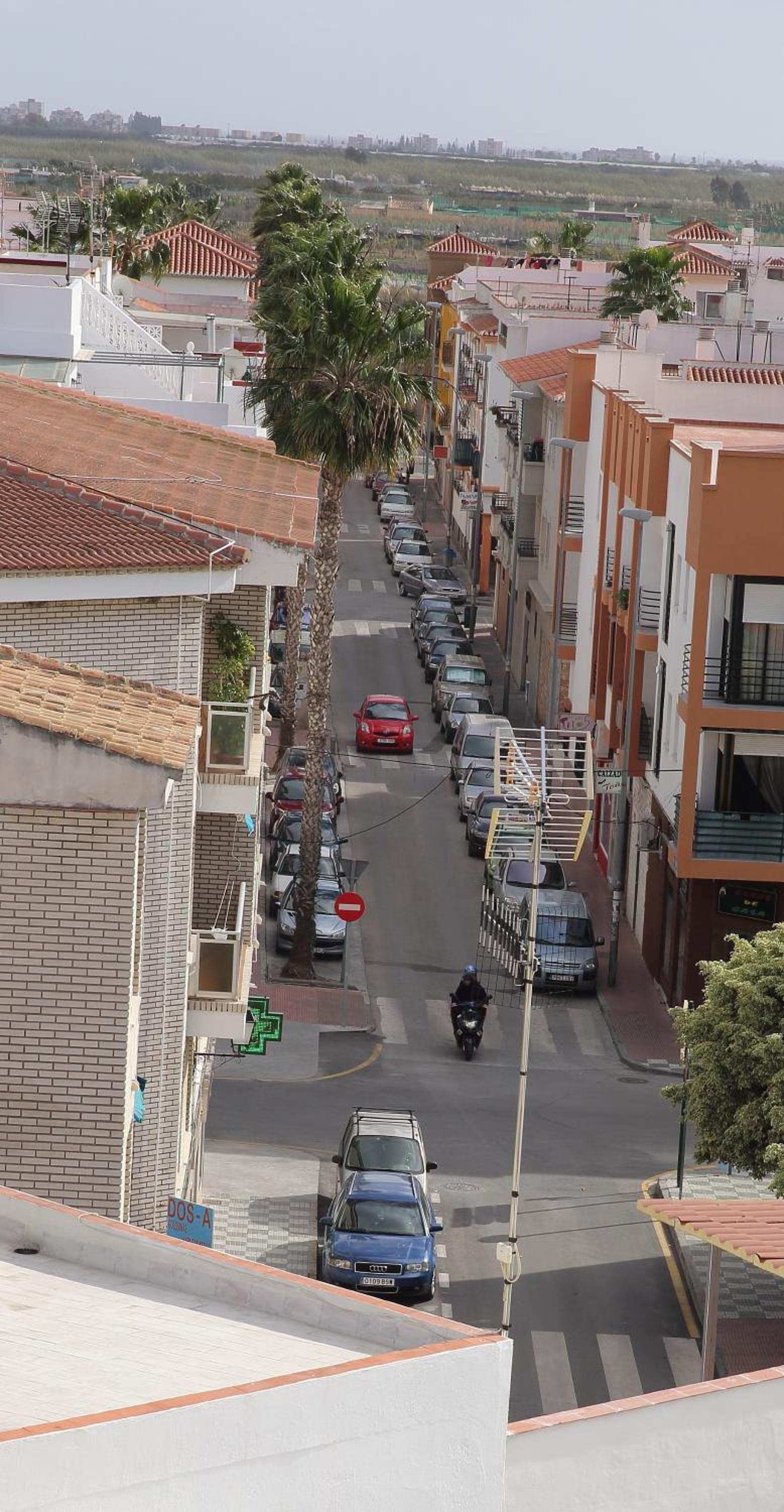 A residential street with parked cars and a motorcyclist. Buildings have red-tiled roofs and balconies.