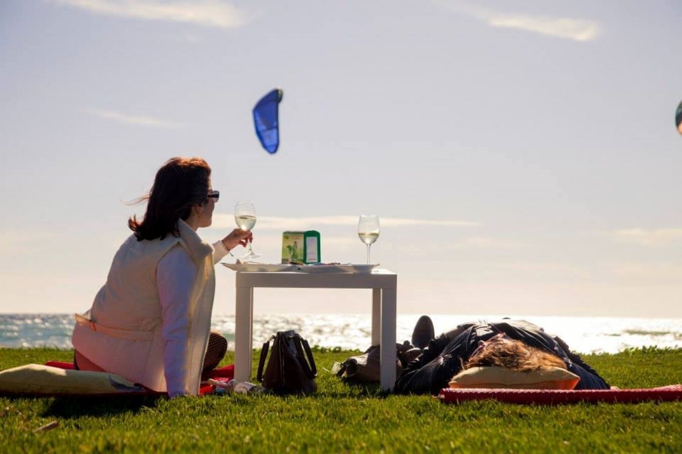 A person sitting on a blanket on grass, holding a glass of wine, with a table in front of them.