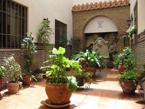 Um pátio com vasos de plantas, piso de azulejos, parede de tijolos e uma fonte de pedra.