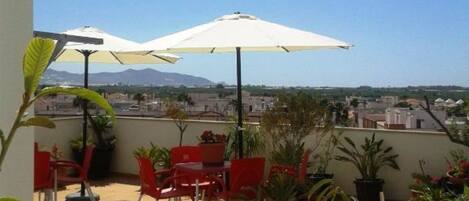 Terraza en la azotea con sillas rojas y sombrillas blancas, rodeada de plantas en macetas y con vistas a montañas lejanas.