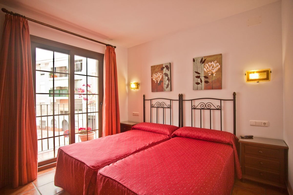 A double bed with red bedding, a window with curtains, and two wall-mounted light fixtures.