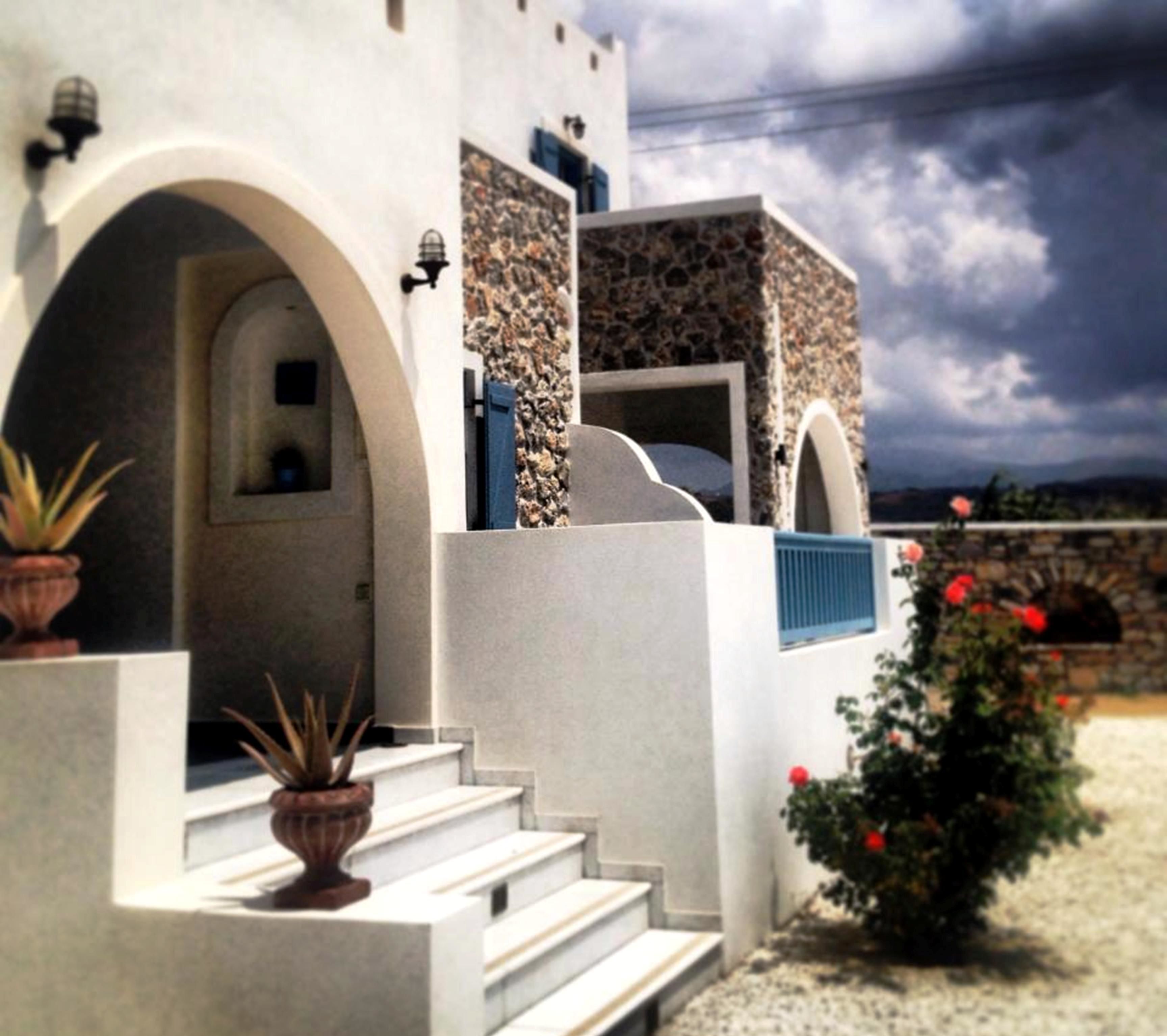 A building with white walls, blue shutters, and a stone facade. There are steps leading to the entrance, and potted plants are placed at the base.