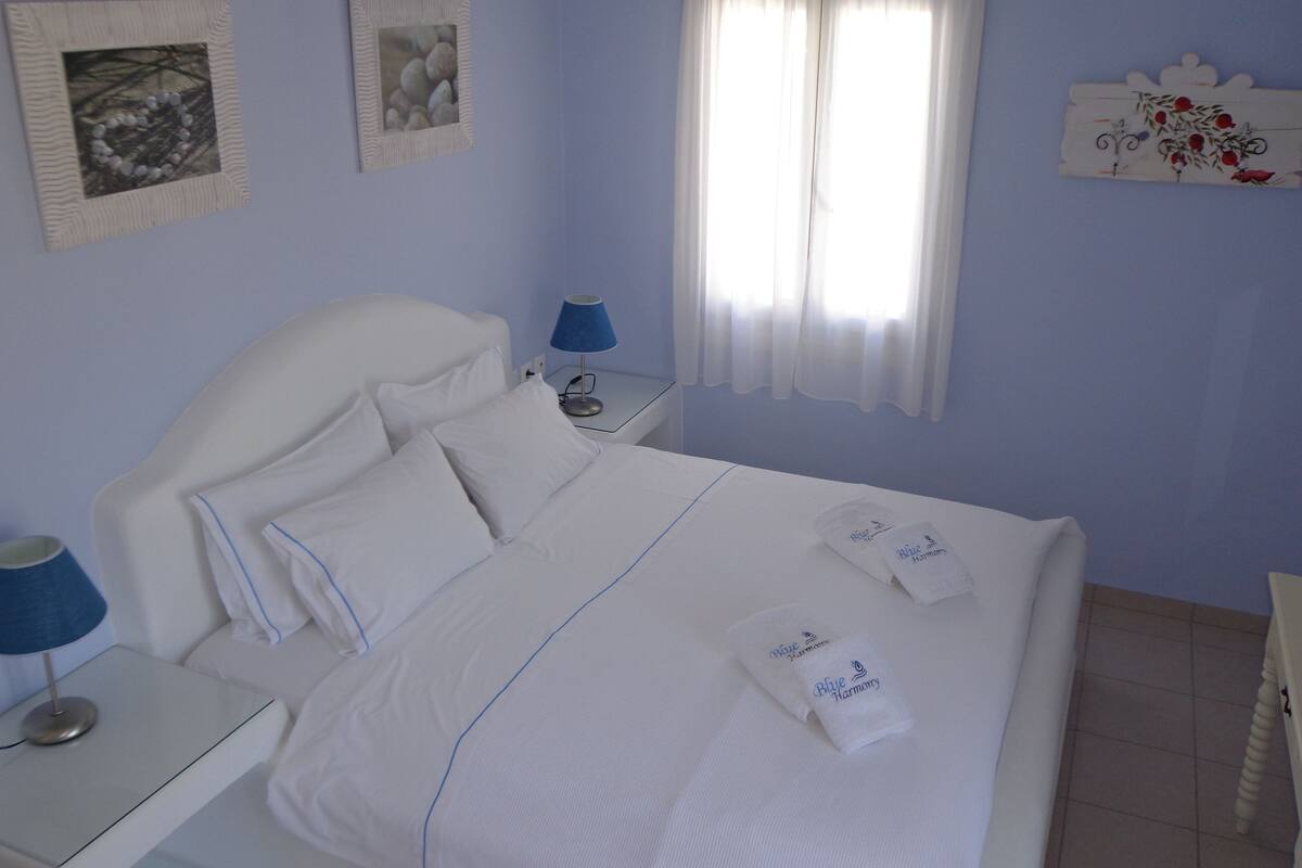 A neatly made bed with white linens and pillows, a bedside table with a lamp, and a wall with framed pictures.