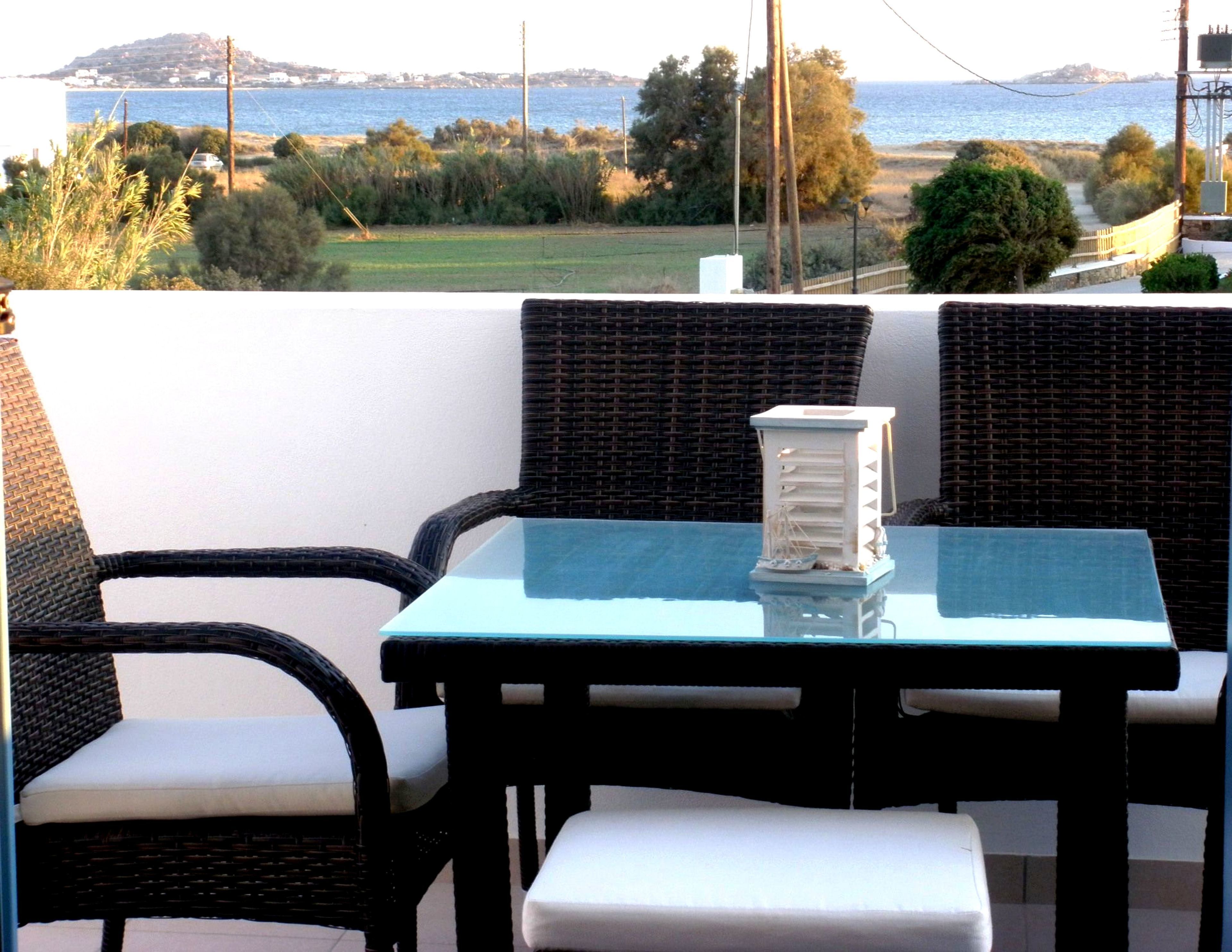 A balcony with wicker furniture, a glass table, and a view of the sea and hills.