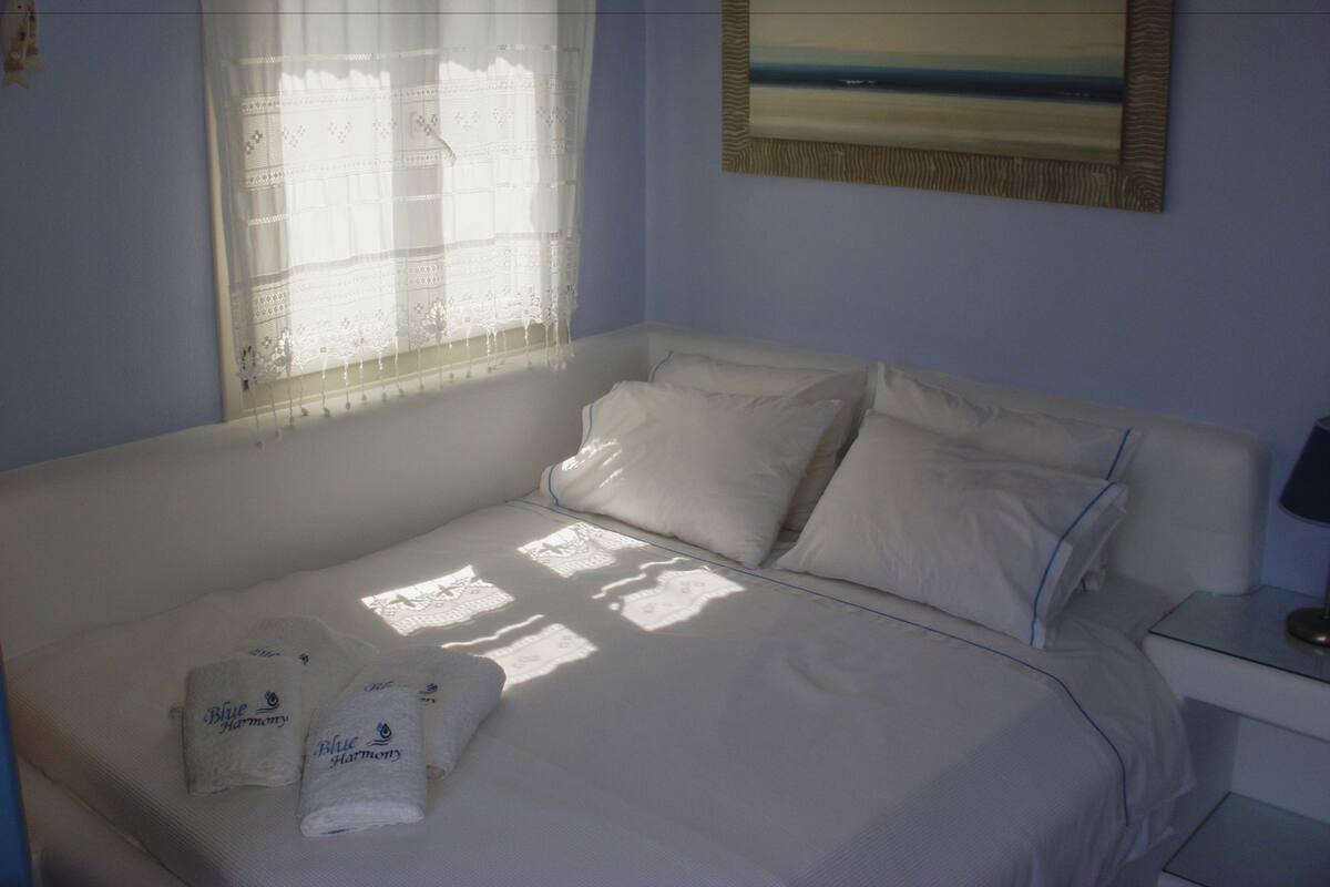 A neatly made bed with white linens and pillows, a window with sheer curtains, and a painting of a beach scene on the wall.