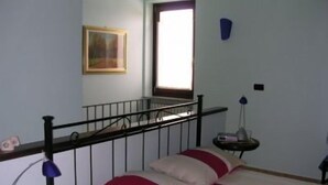 A bedroom with a bed, a window, a painting on the wall, and a small table with a lamp. - L'Angolo di Rosina (Novello)