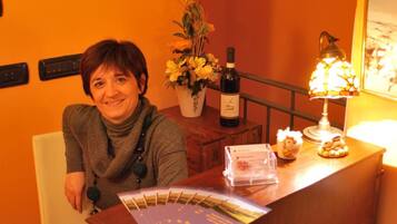 Una donna seduta a una scrivania di legno in una stanza illuminata con calore, con una parete arancione, un vaso di fiori, una bottiglia di vino e una lampada.