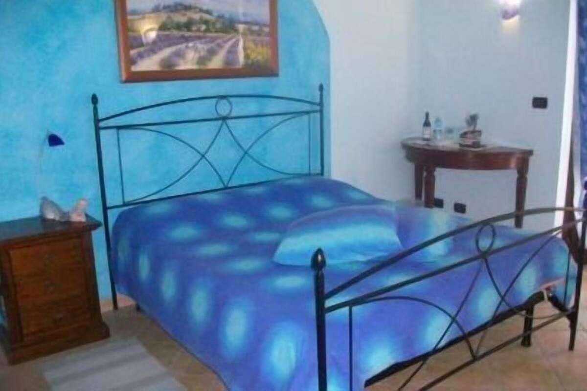 A bedroom with a bed, a dresser, a painting on the wall, and a small table with a bottle.