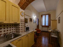 Una cucina compatta con mobili in legno, un forno a microonde e un lavandino. Si vede un orologio al muro e una finestra con tende.