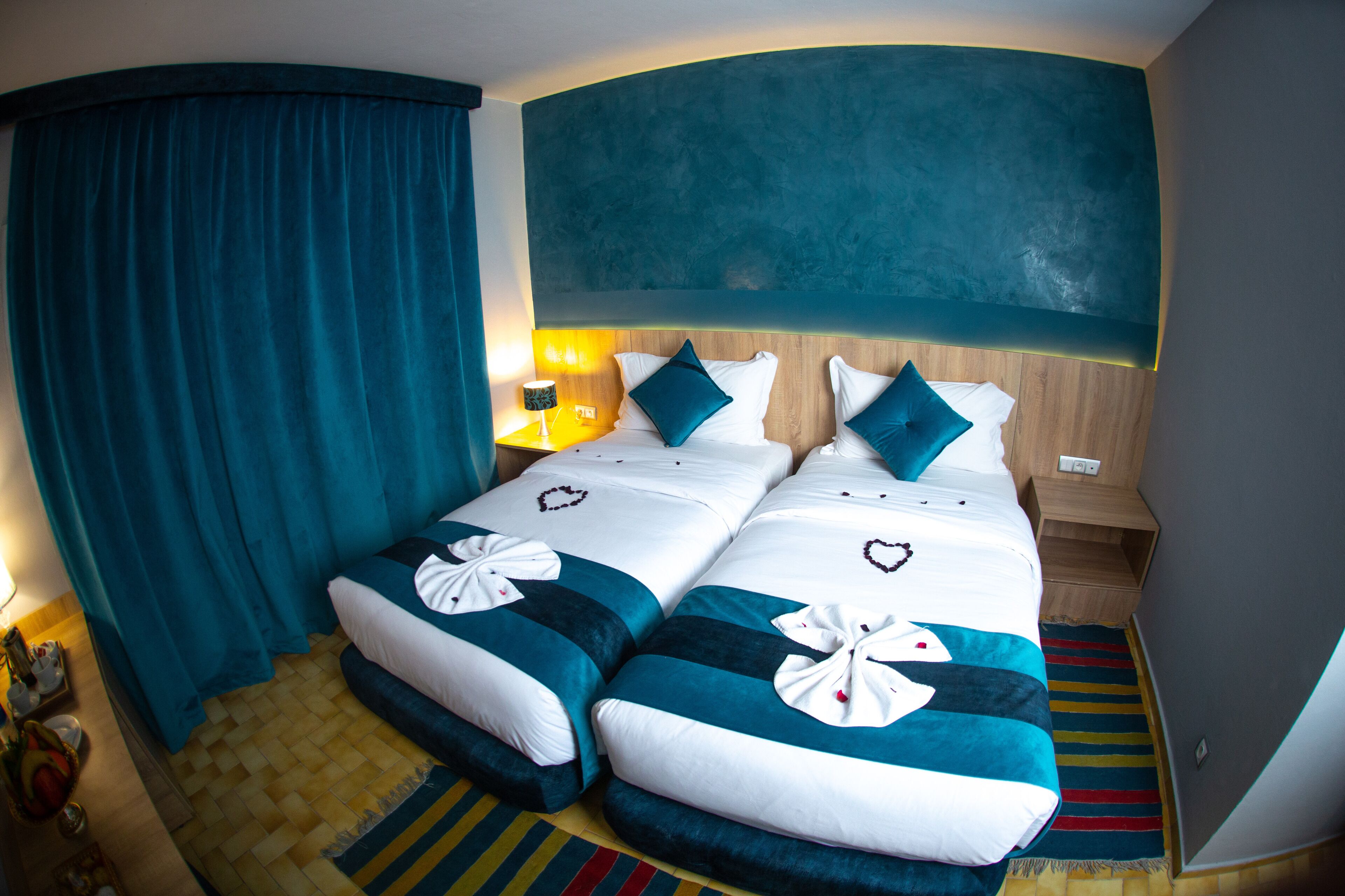 A hotel room with two beds, teal and white bedding, a wooden headboard, a nightstand with a lamp, and a colorful striped rug.