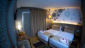 Habitación de hotel con dos camas, un escritorio con frutero, televisión y un detalle decorativo en la pared.