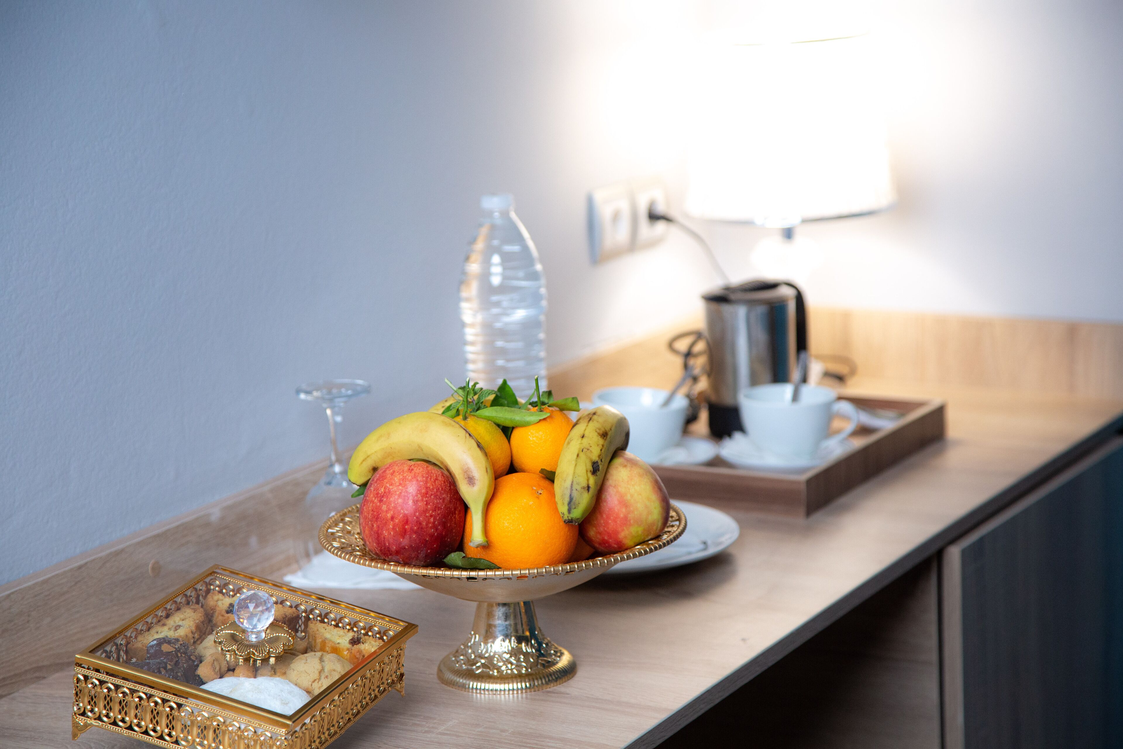 A breakfast setup with a fruit bowl, a bottle of water, a coffee pot, and a tray with cookies.