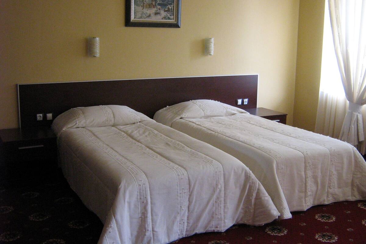 A hotel room with two single beds, a nightstand, a framed picture on the wall, and a window with curtains.
