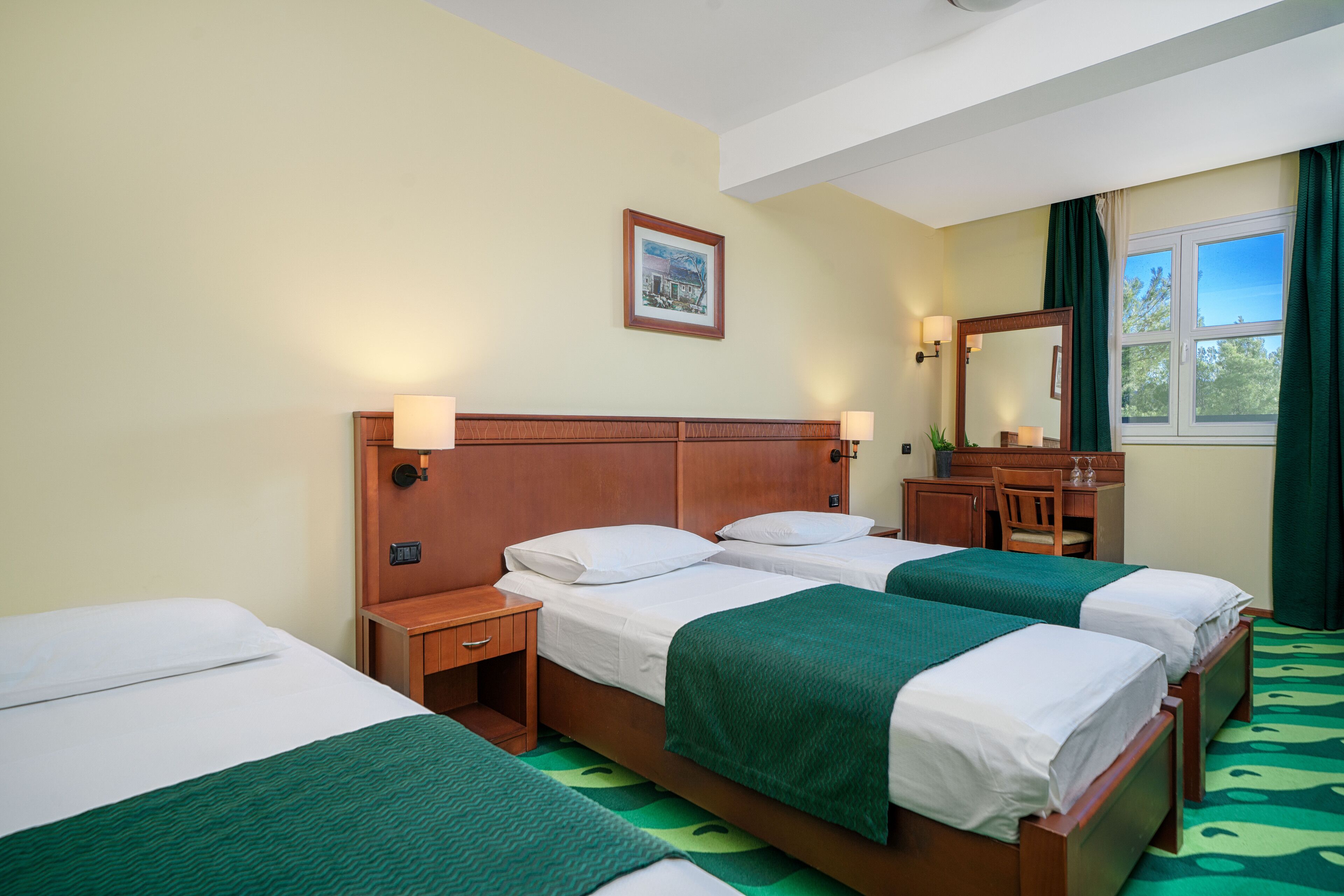 A hotel room with two single beds, a wooden headboard, a small desk with a chair, a mirror, and a window with curtains.