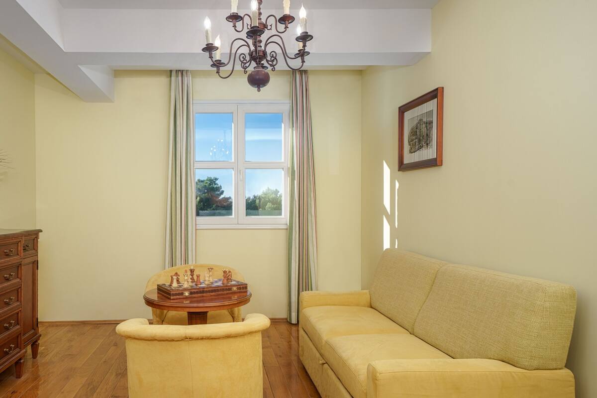Ein Wohnzimmer mit gelbem Sofa, Holzschrank, Schachbrett und einem Fenster mit Vorhängen.