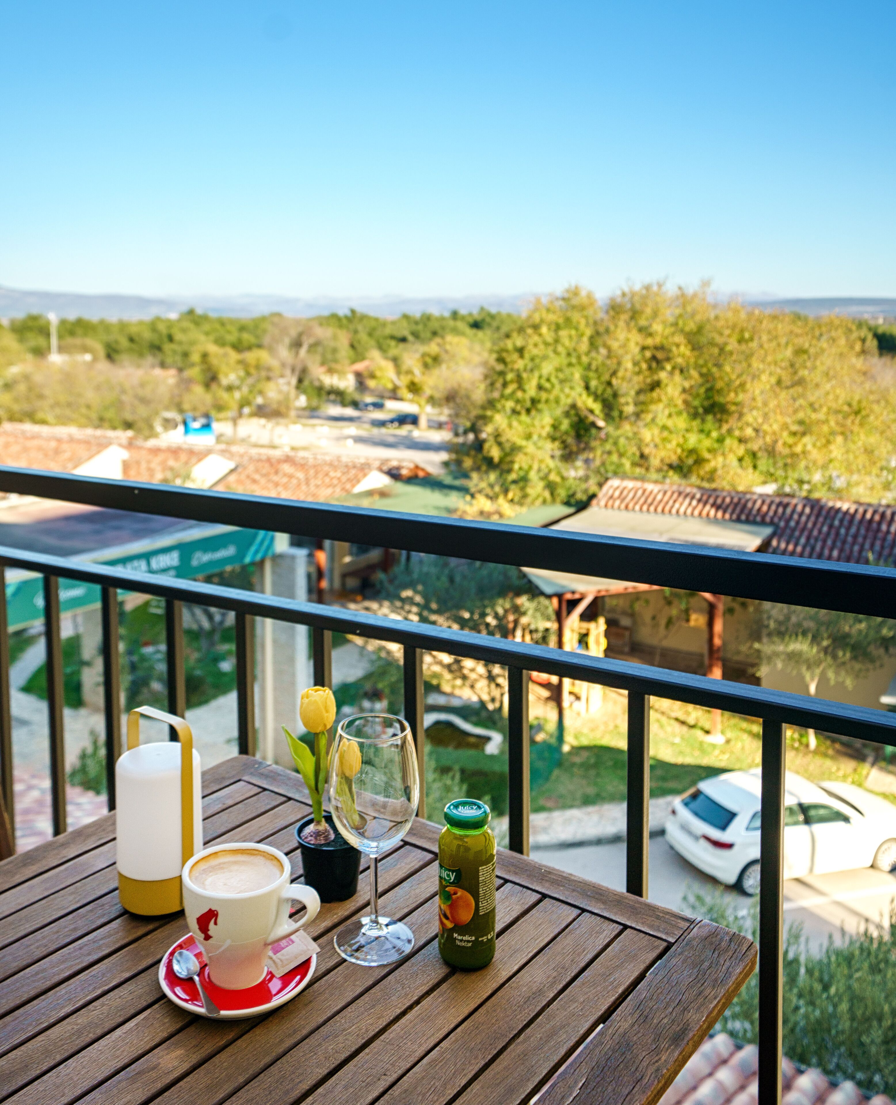 Una mesa de madera con una taza, un vaso y una botella en un balcón con vistas a una zona residencial.
