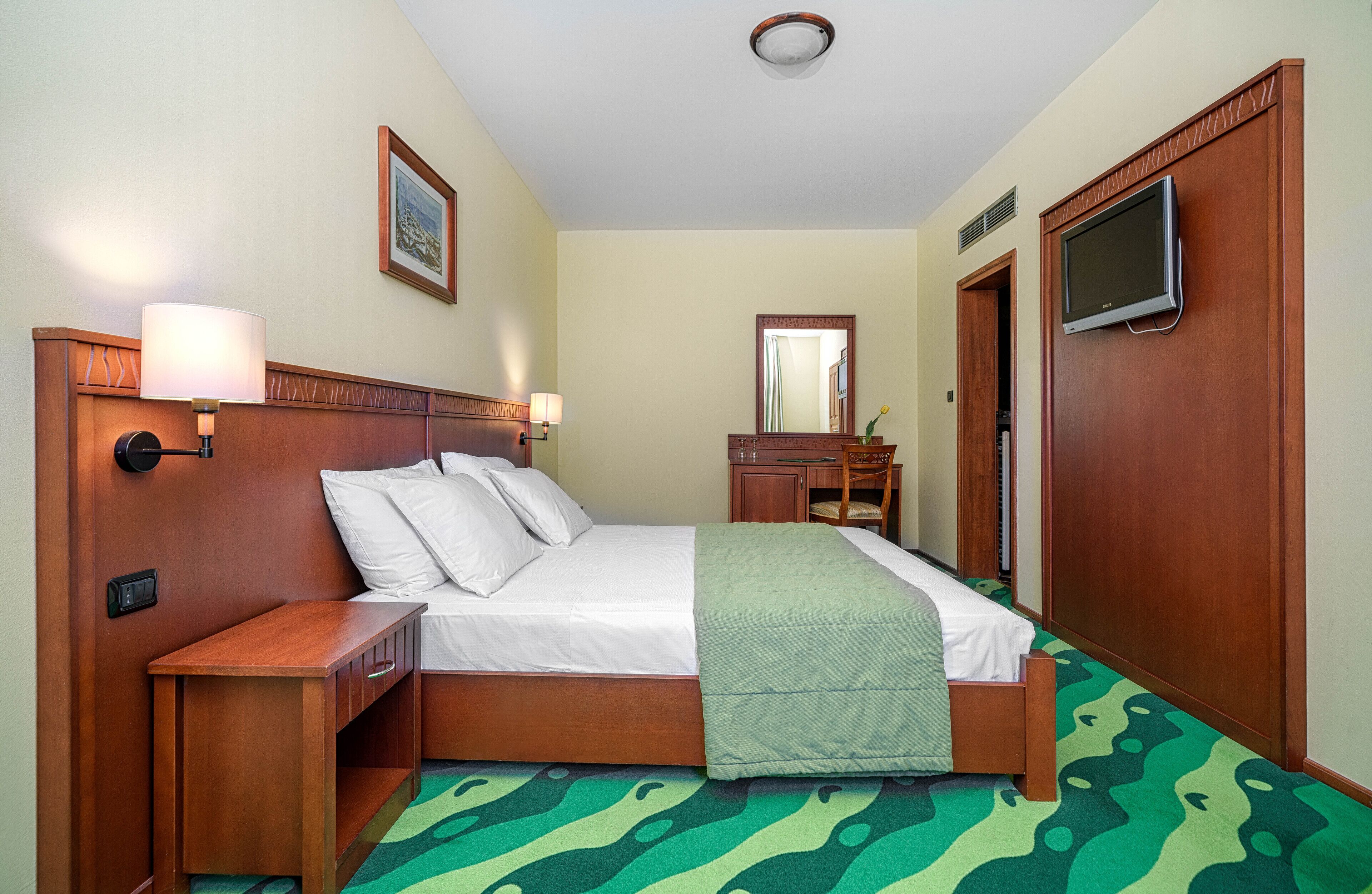 A hotel room with a wooden bed, bedside tables, a TV, a mirror, and a green patterned carpet.