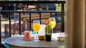 Una mesa con una taza, un vaso de jugo de naranja, una tulipa y un libro en un balcón con vistas a árboles.