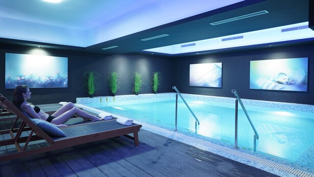 Un área de piscina interior moderna con una tumbona, una piscina y pantallas en la pared que muestran diversas imágenes.