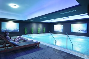 Un espace piscine intérieur moderne, doté d’un coin salon avec des fauteuils, d’une piscine et d’écrans fixés au mur diffusant diverses images.