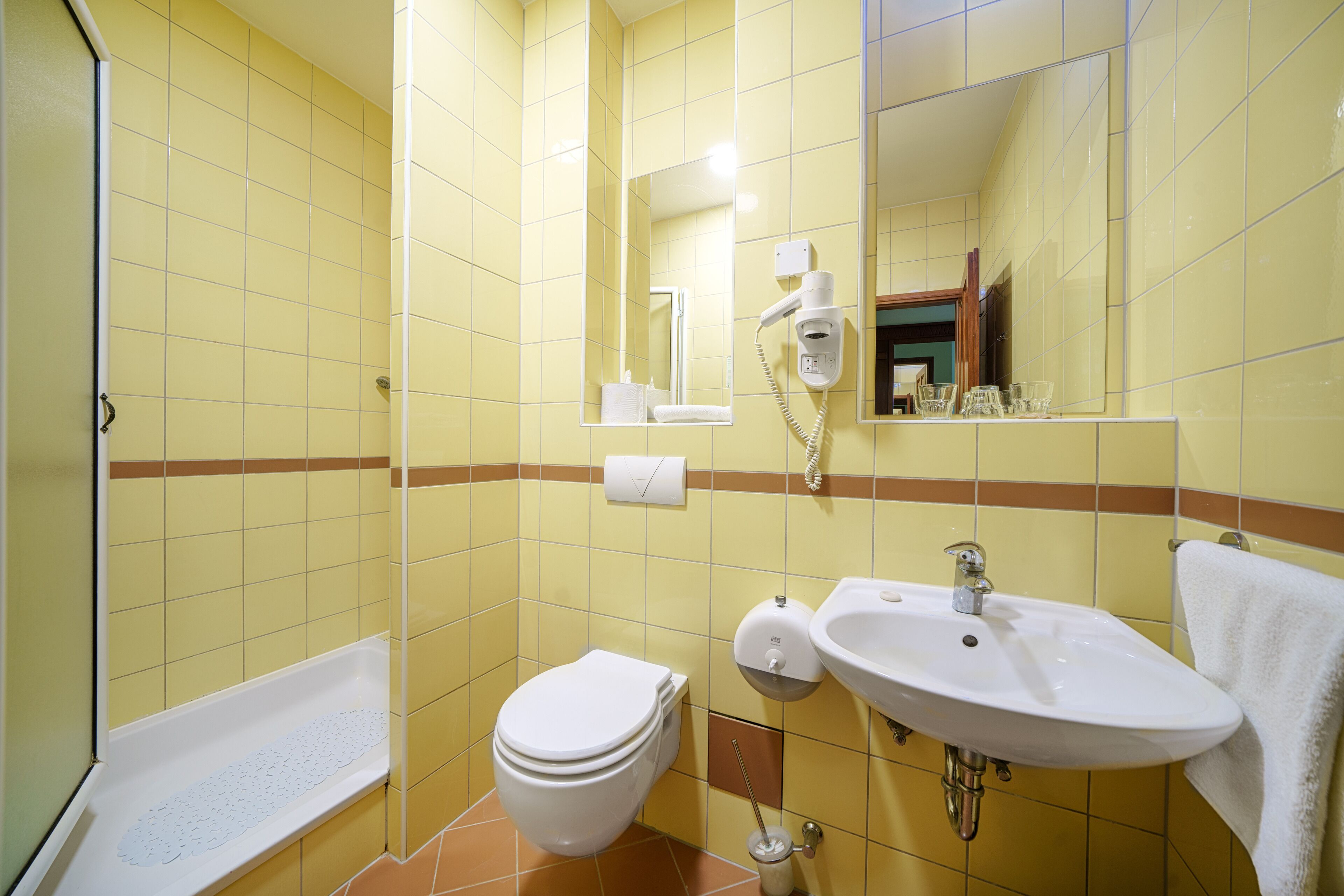 Une salle de bain avec des carreaux jaunes, des toilettes blanches, un lavabo, un miroir et une douche.