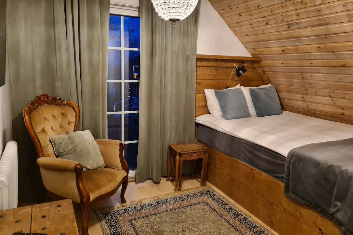 A cozy room with a wooden headboard bed, a plush armchair, a small wooden nightstand, and a large window with curtains.