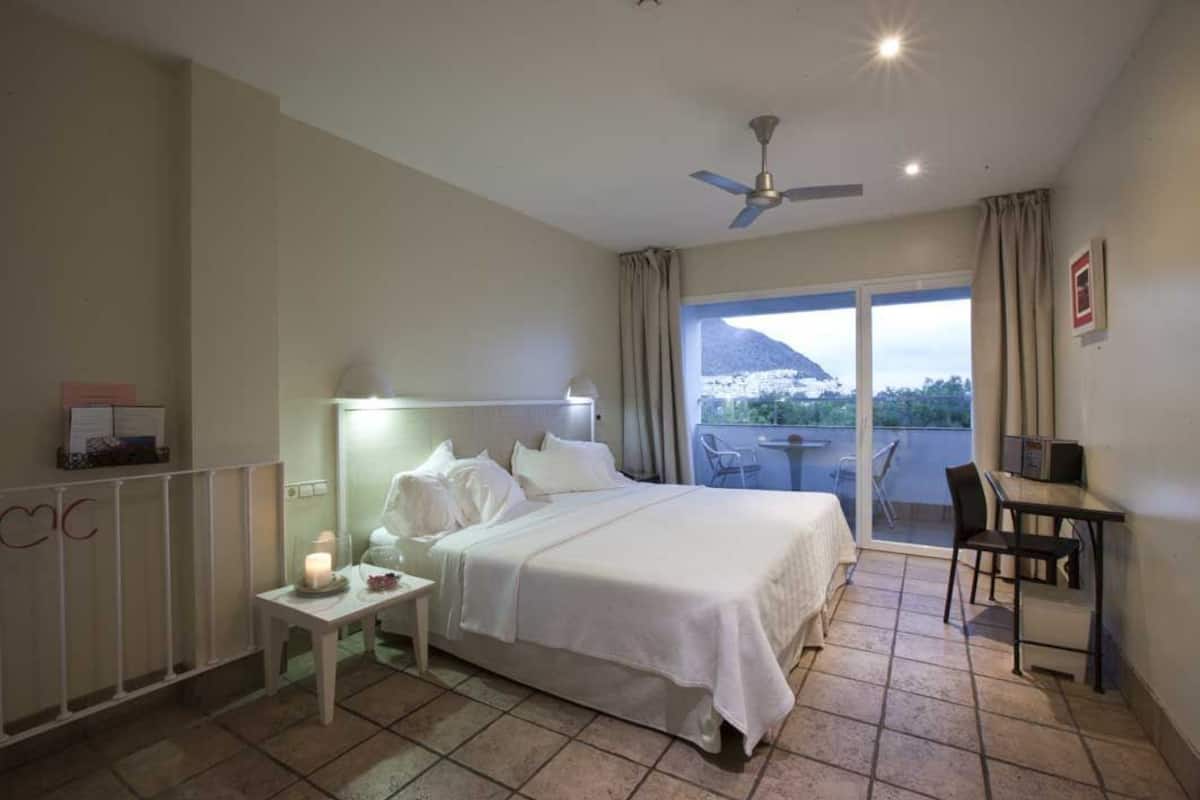 A hotel room with a bed, bedside table, lamp, and a view of a mountain through a large window.