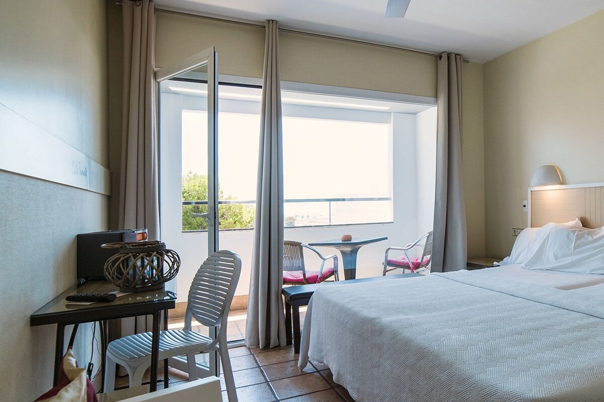 Quarto de hotel com cama, escrivaninha com computador, cadeira, mesinha e varanda com vista.