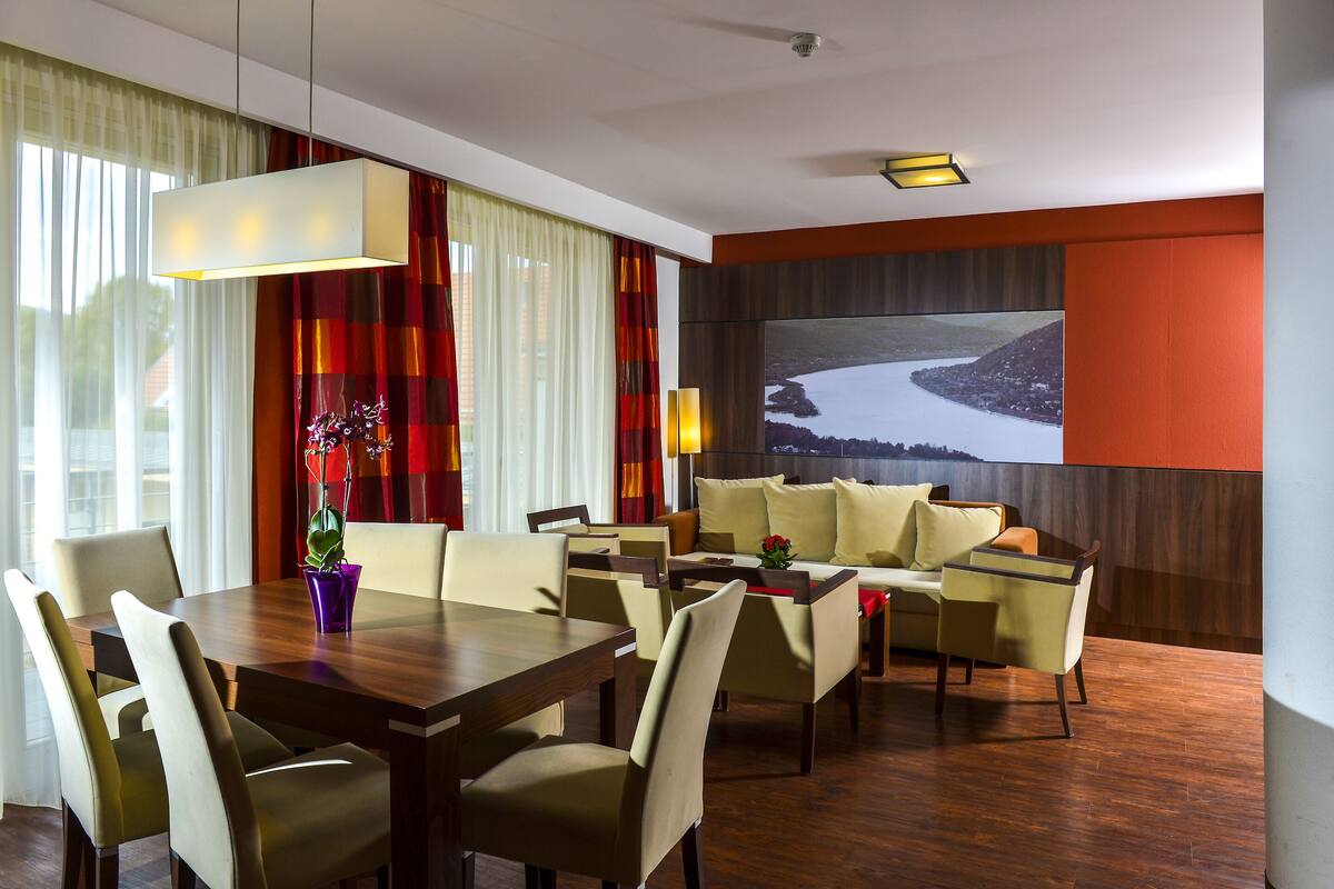 A hotel room with a dining area, a sofa set, and a large window with curtains.