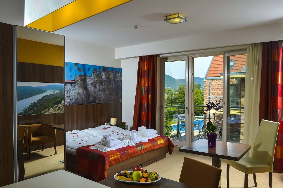 A hotel room with a bed, a dining table, a chair, a fruit bowl, a painting, and a balcony with a pool view.