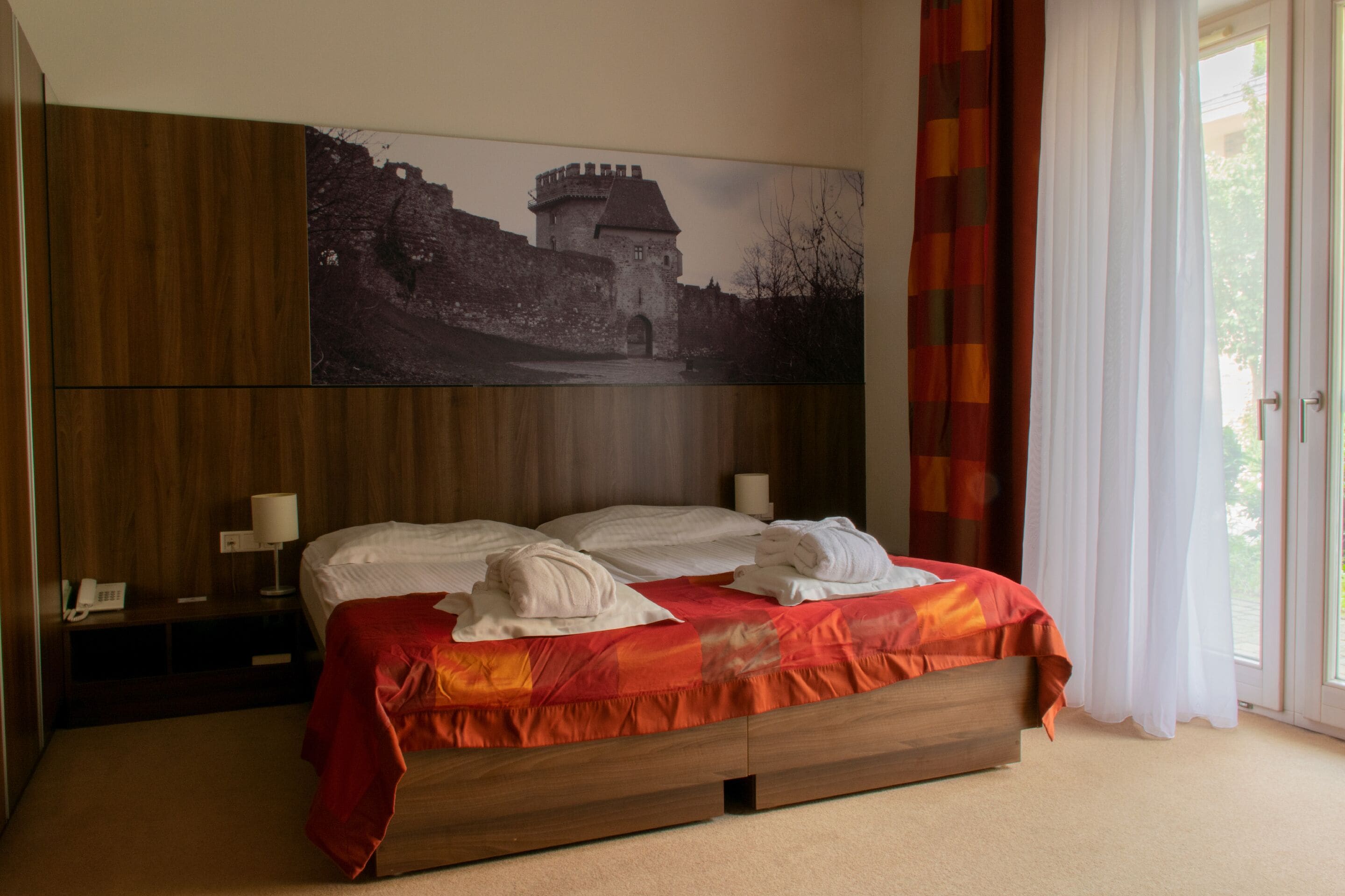Una habitación de hotel con una cama grande, cabecero de madera, un cuadro colgado de un castillo, una mesita de noche con una lámpara y una ventana con cortinas.