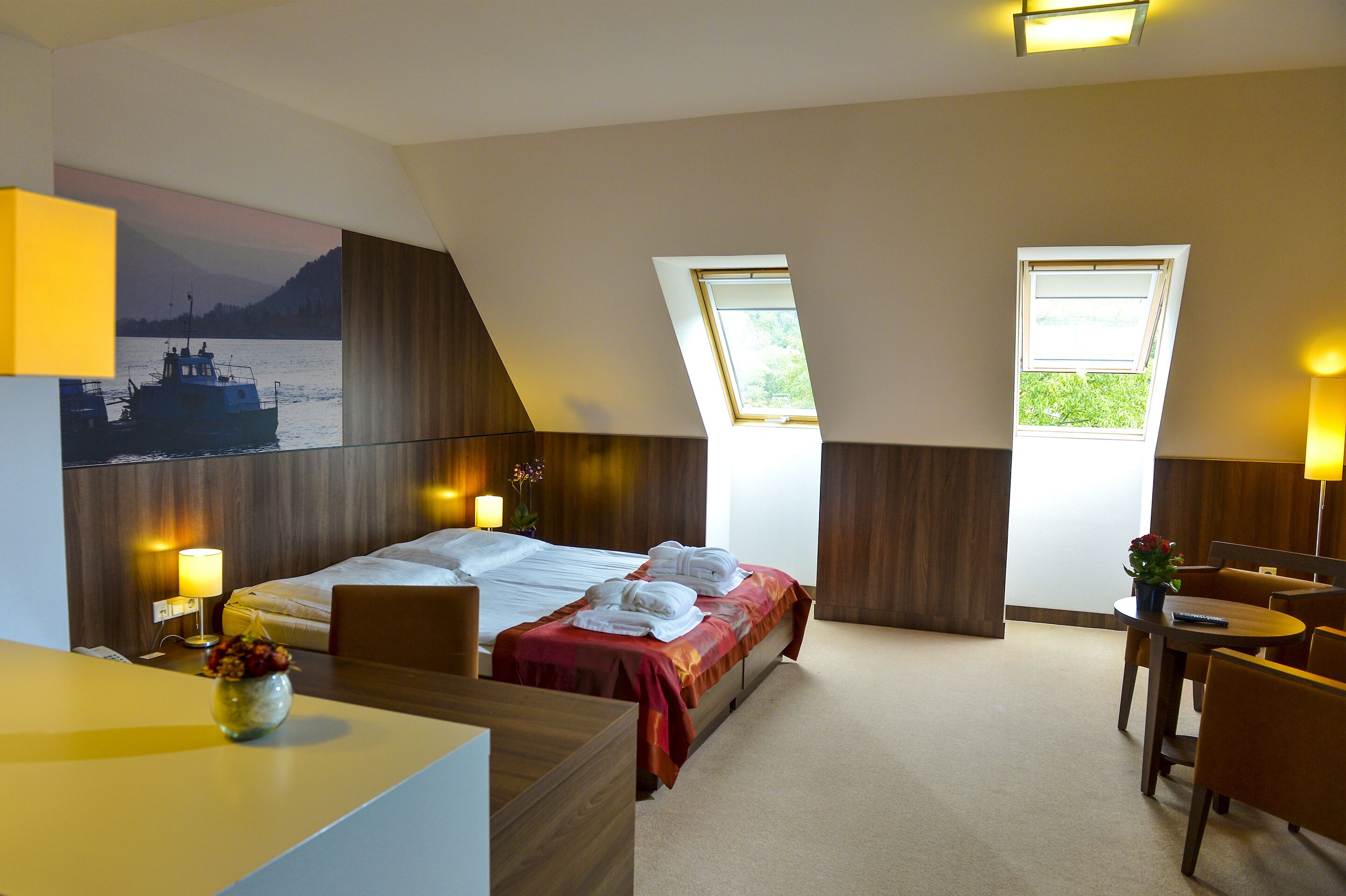 A hotel room with a bed, a desk, a chair, a small table, and two windows.