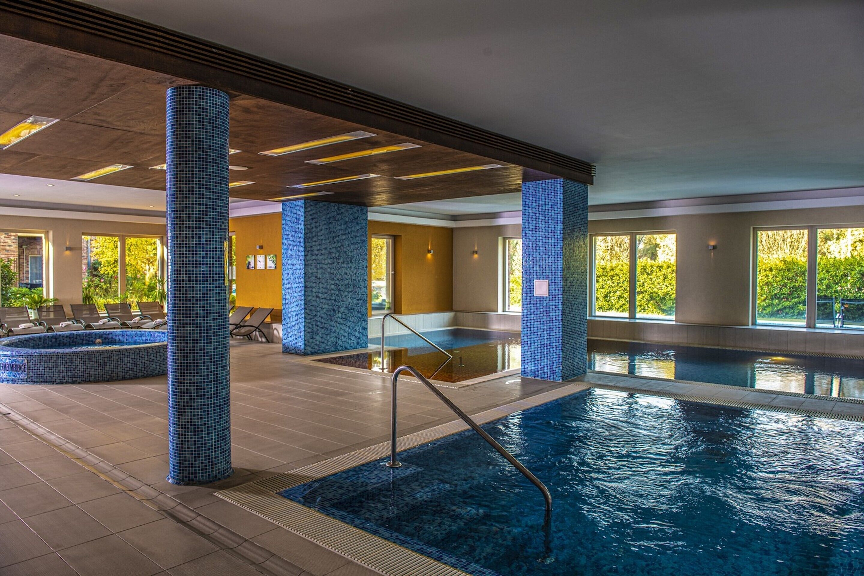 Une piscine intérieure aux murs et aux piliers recouverts de carrelage, un espace baigné de lumière avec des chaises longues, et une vue dégagée sur l’extérieur.