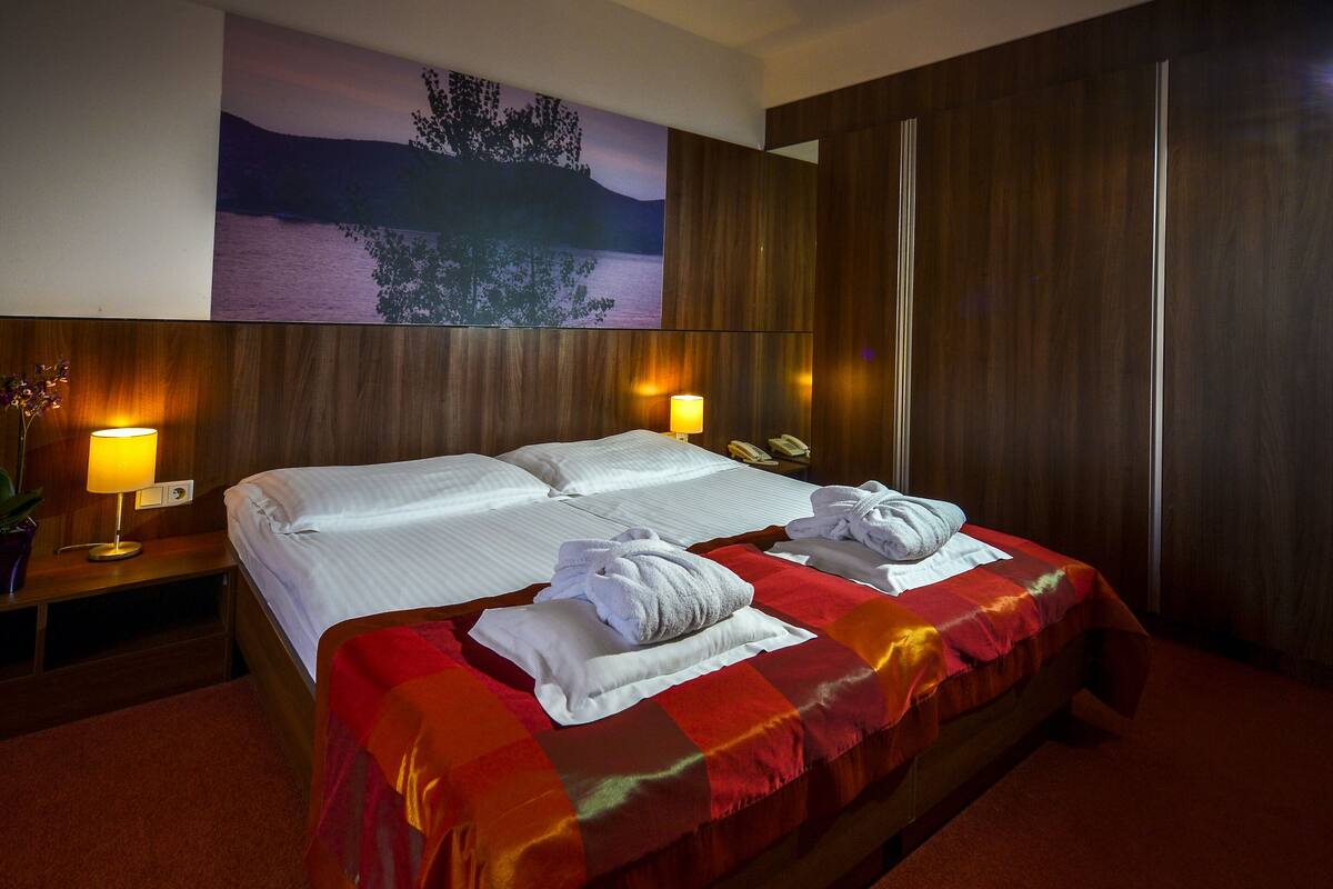 A hotel room with a large bed, two bedside lamps, a nightstand with a lamp, a telephone, and a painting of a landscape on the wall.