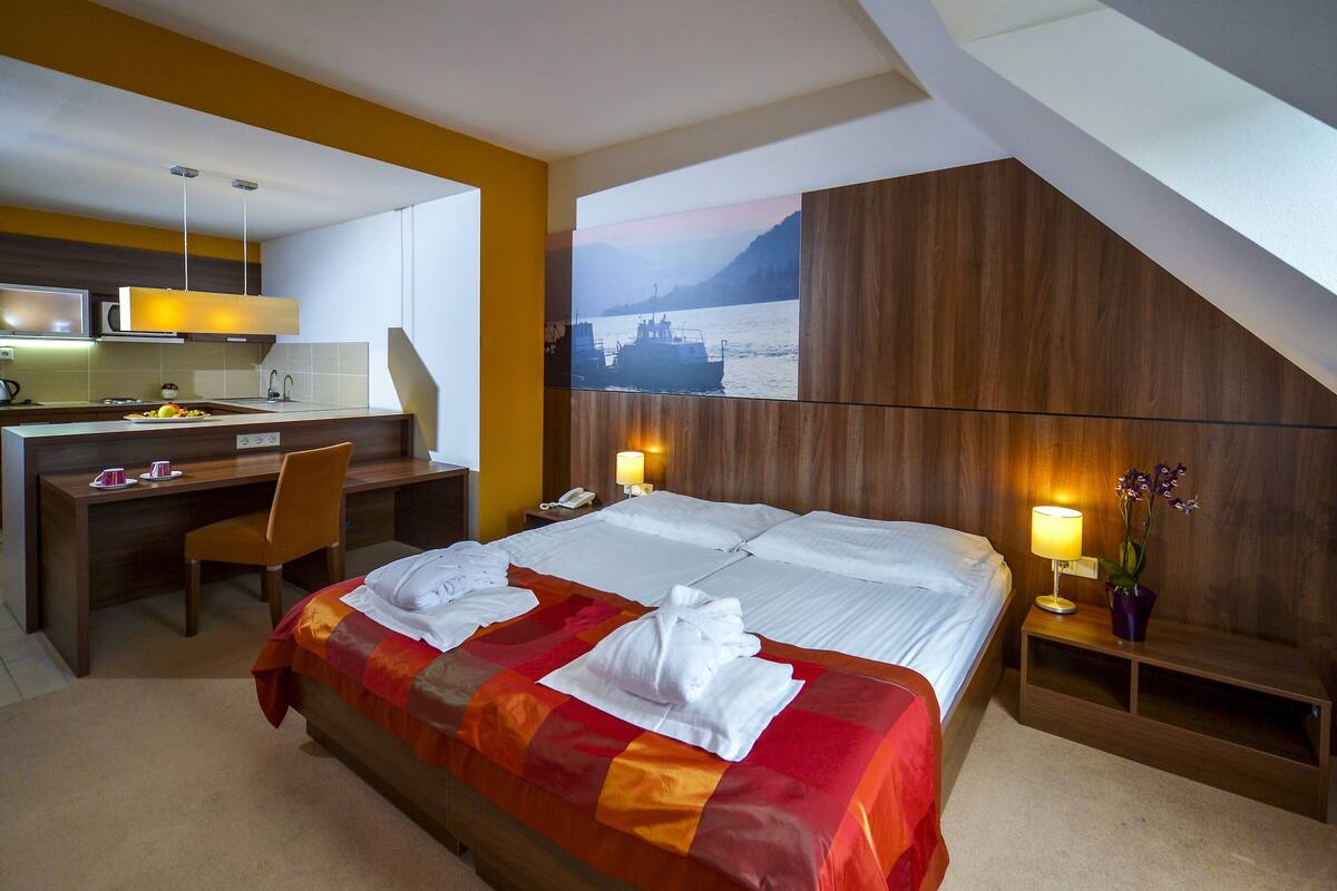 A hotel room with a bed, desk, chair, bedside table, lamp, and a painting of a boat on water.
