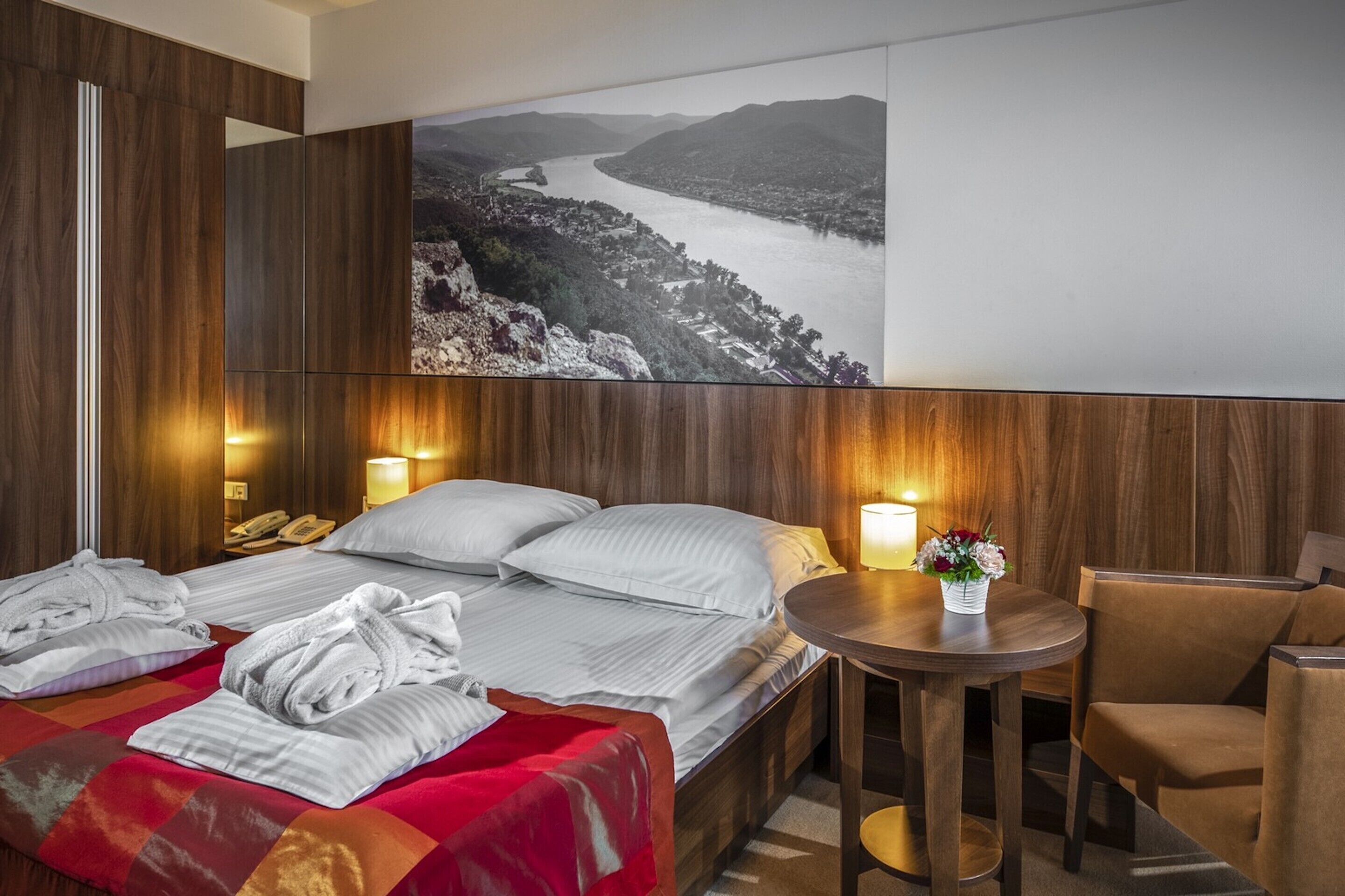 A hotel room with a large bed, two bedside tables, a small round table with a vase of flowers, and a large framed picture of a landscape on the wall.