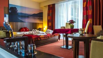 A hotel room with a bed, a dining table, a chair, a lamp, and a painting on the wall.