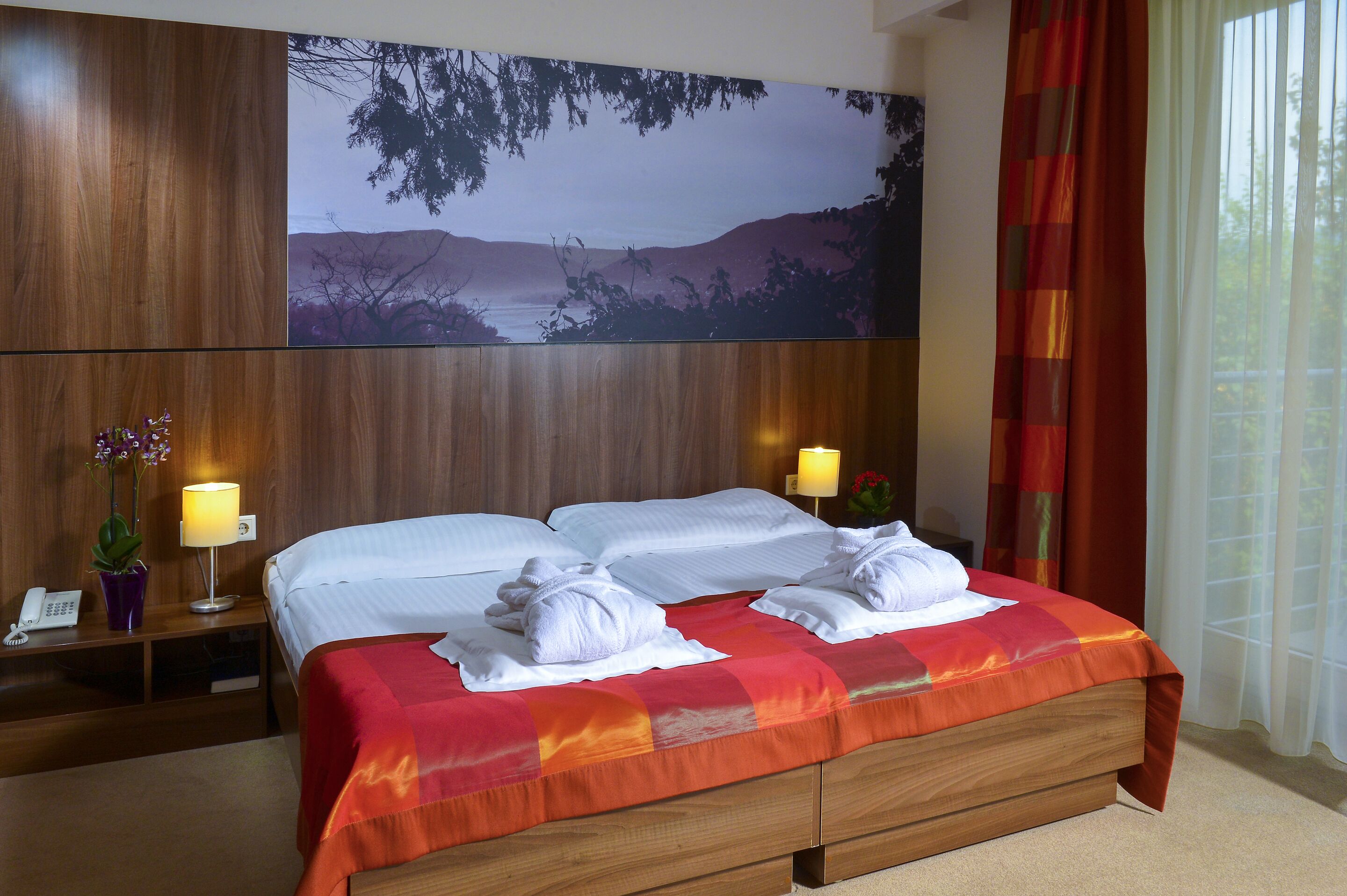 Une chambre d’hôtel avec un grand lit, deux tables de chevet, une petite table d’appoint, une lampe et un grand tableau représentant un paysage accroché au mur.