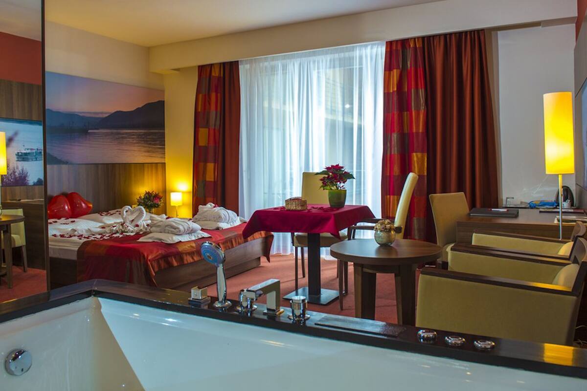 A hotel room with a large bed, a desk, a chair, a table with a vase, and a large window with curtains.