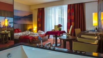 A hotel room with a large bed, a desk, a chair, a table with a vase, and a large window with curtains.