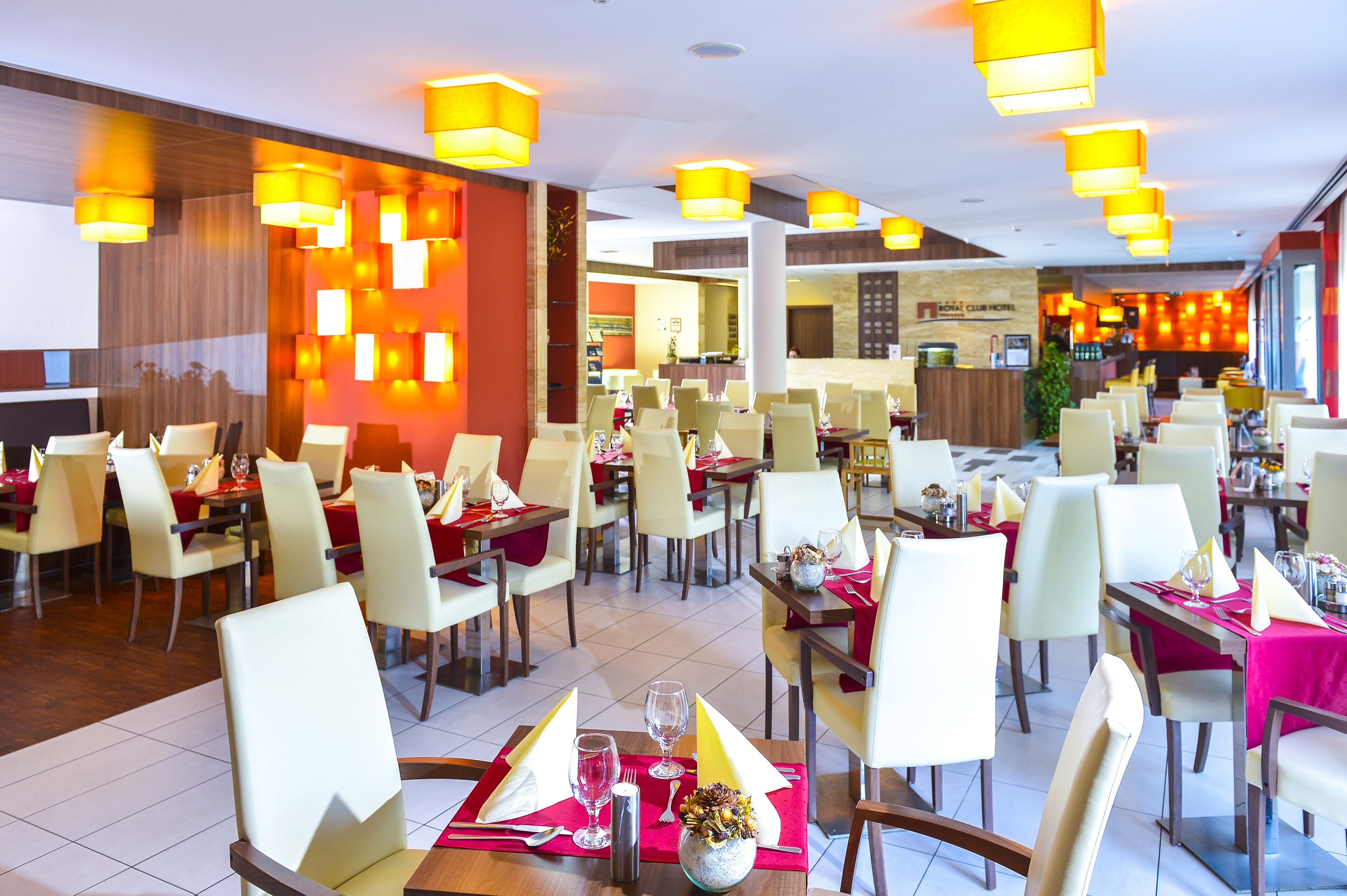 A hotel dining area with multiple tables and chairs, bright overhead lighting, and a visible reception desk in the background.
